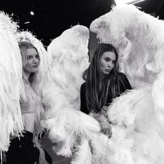 two women standing next to each other in front of white angel wings on display at a fashion show