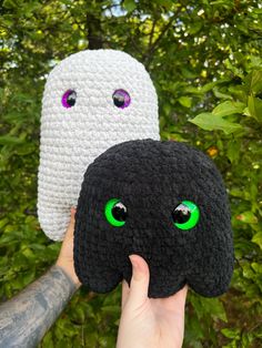 two crocheted objects with green eyes are held up in front of some trees