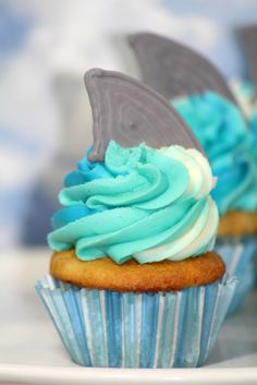 two cupcakes with blue frosting and a shark fin on top are sitting on a plate