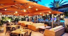 an outdoor dining area with tables and chairs under umbrellas at night time, surrounded by palm trees