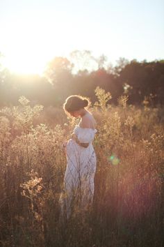 a pregnant woman standing in tall grass with the sun shining behind her and looking down at her belly