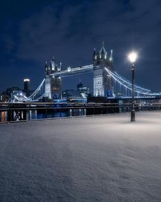 the tower bridge is lit up at night with snow on the ground and lights in the background