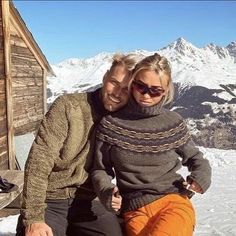 a man and woman sitting on top of a wooden bench in front of snow covered mountains