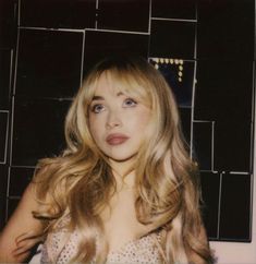 a woman with long blonde hair standing in front of a tiled wall and looking at the camera