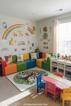 Brightly colored children's playroom featuring a rainbow mural, animal decals, and a cozy seating area with colorful foam blocks. The room includes a tree-patterned rug, a small table with chairs, and a craft station with organized supplies, creating a vibrant and inviting space for kids to play and learn. How To Create A Play Area In Living Room, Kids Stage In Playroom, Infant Play Area In Living Room, Preschool Room Ideas, Childminders Playroom Home Daycare