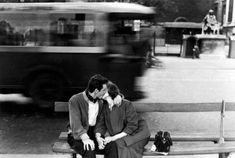 black and white photograph of two people sitting on a bench in front of a bus