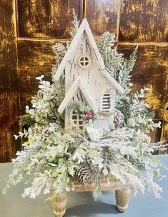 a white birdhouse is surrounded by greenery and pine cones on a wooden table