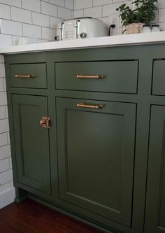 a green kitchen cabinet with brass handles and knobs on the doors, in front of a white tiled wall