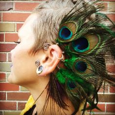 a woman with peacock feathers on her ear and piercings in front of a brick wall