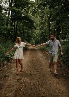 a man and woman holding hands while walking down a dirt road in the middle of a forest