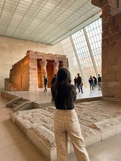 a woman standing in front of an ancient building with people walking around it and looking up at the sky