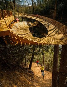 two people riding bikes on a dirt path in the woods with a wooden structure above them