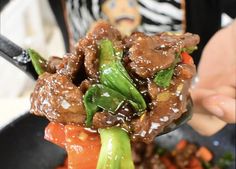 a person holding up a spoon full of stir fry meat and vegetables with sesame seeds