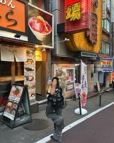 a woman standing on the side of a street talking on a cell phone in front of a restaurant