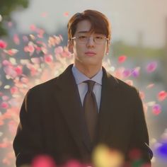 a young man wearing glasses and a suit with flowers in the back ground behind him