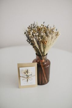 dried flowers in a vase next to a card on a white table top with a brown frame