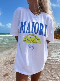 a woman standing on the beach wearing a white shirt with blue and yellow lettering that says major