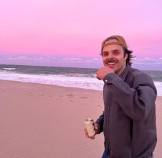 a man standing on top of a sandy beach next to the ocean holding a cup