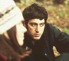 a man and woman sitting next to each other in the woods looking at each other