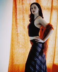 a woman standing in front of a curtain with her hands on her hips, wearing a black top and plaid skirt