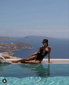 a woman in a black swimsuit sitting on the edge of a pool next to an ocean