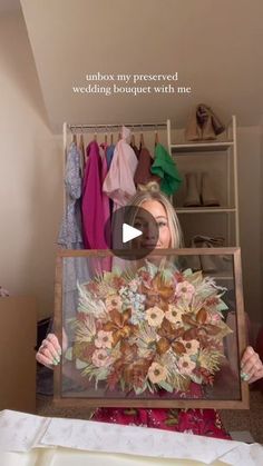 a woman holding up a painting with flowers on it in front of a closet full of clothes