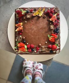 a person standing in front of a chocolate cake with fruit on it and berries around the edges