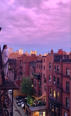 a woman standing on top of a balcony next to tall buildings at dusk with city lights in the background