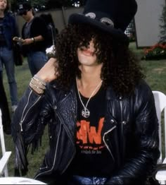 a man with long hair wearing a black hat and leather jacket sitting on a white chair