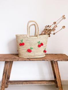 a straw bag with cherries on it sitting on a wooden bench next to flowers