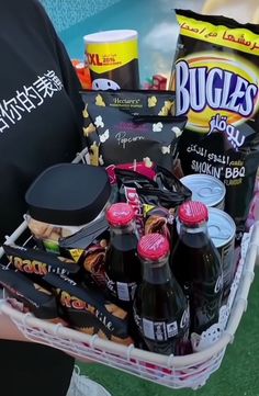 a person holding a basket full of snacks and drinks on the grass with other items in it