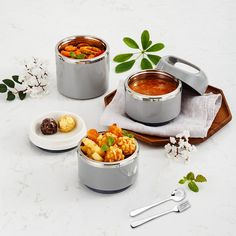 three silver pots with food in them on a white counter top next to flowers and spoons