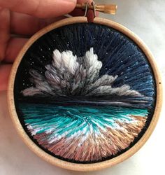 a hand holding a small embroidery art piece with an ocean scene in the center and clouds above it