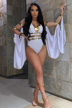 a woman in a white bodysuit and gold jewelry standing next to a stone wall