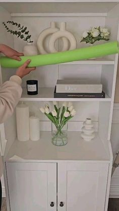 a person holding a green object over a white cabinet with flowers on it and bookshelves