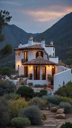 a white house in the middle of some bushes and trees with mountains in the background