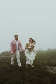 a bride and groom walking through the foggy grass on top of a hill holding hands