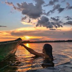 a woman is in the water holding onto a boat at sunset with clouds above her