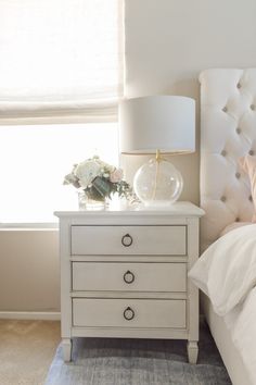 a white nightstand with flowers on top and a lamp next to it in front of a window