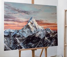 an easel with a painting on it in front of a mountain range at sunset