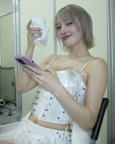 a woman in white dress holding a cell phone and looking at her reflection on the mirror