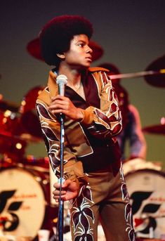 a man with an afro holding a microphone in front of drum set and drums behind him