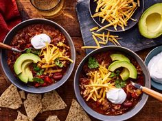 two bowls filled with chili and cheese on top of a blue wooden table next to silver spoons