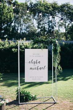 a welcome sign in the grass with greenery around it and hanging from a wire