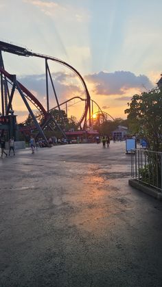 the sun is setting behind an amusement park roller coaster at dusk with people walking around