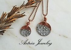 two silver and copper pendants on a white marble surface with dry leaves in the foreground