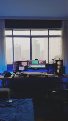 there is a desk with two monitors and speakers in front of a window that looks out onto the city