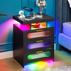 a night stand with three drawers and colorful lights on the bottom, in front of a blue wall