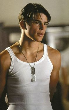 a young man wearing a white tank top with a cross necklace on his neck, looking at the camera