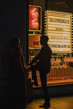 a man and woman standing in front of a movie theater at night with lights on the marquee